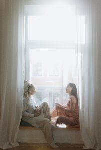 Two woman sitting in window seat with mugs talking