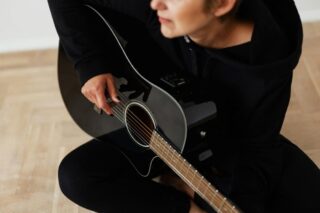 Person sitting cross-legged on floor in black long sleep shirt playing acoustic guitar