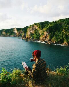 Person sitting in grass next to water with a book and a teacup