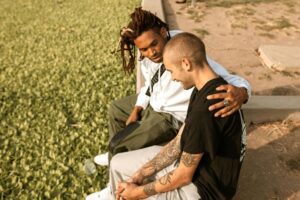 Two men sitting on a concrete wall one with arm around the other talking